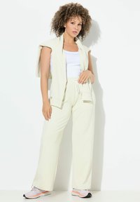 Lichtcrème-kleurige athleisure-outfit bestaande uit een mouwloos topje en losse broeken met een elastische tailleband. Draagt bijpassende grijze en oranje sneakers.