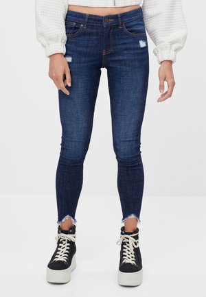 Jean skinny bleu foncé avec une taille mi-haute, léger vieillissement sur le devant et ourlets effilochés, associés à des baskets montantes noires.