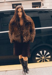 Braune, zottelige Kunstpelzjacke, kniehoher Wildlederrock und schwarze, hochhackige Lederstiefel. Im Hintergrund sind ein schwarzes Auto und Pflastersteine zu sehen.