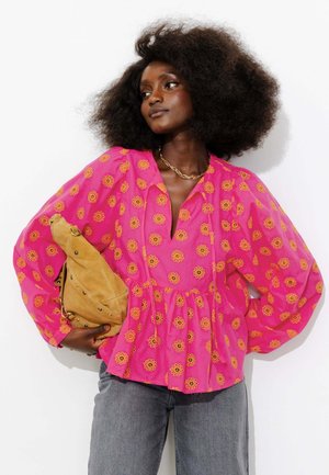 Femme aux cheveux bouclés et volumineux porte un chemisier rose à motifs de fleurs jaunes, un jean gris, un collier en or et tient un sac en daim beige.