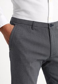 Muž nosící texturované tmavě šedé kalhoty a bílou košili s dlouhým rukávem, s jednou rukou v kapse.