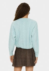 Kvinna med lockigt hår som bär silverfärgade hoop örhängen, en ljusblå kablage stickad tröja och en brun rutig kjol, sedd bakifrån.