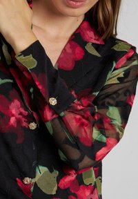 Blusa de manga larga en tela negra con un patrón floral en rojo y verde. Presenta mangas transparents y detalles en oro en los botones con un acabado texturizado.