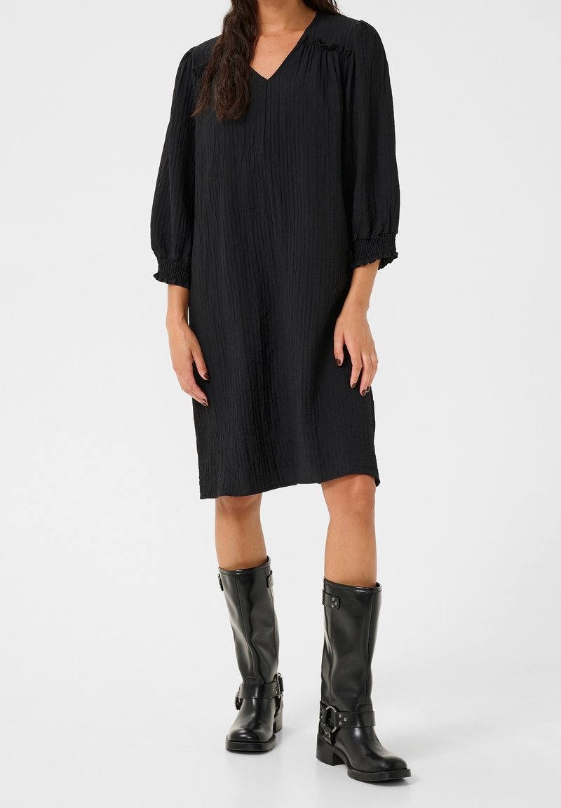 Femme portant une robe noire texturée longueur genoux avec des manches trois-quarts bouffantes et des bottes en cuir noir montantes jusqu'au genou ornées de détails à boucles.