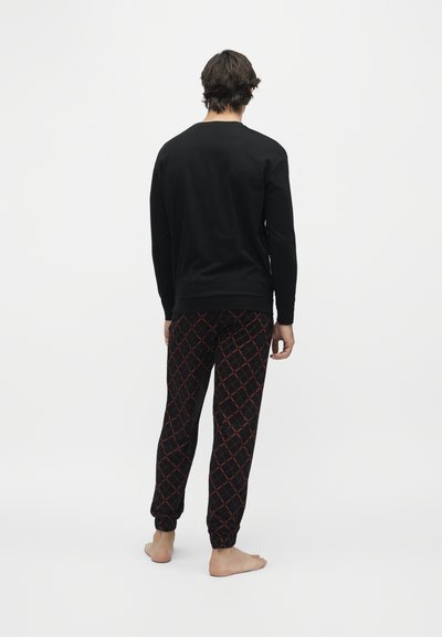 Homme debout pieds nus, de dos, portant un haut à manches longues noir et un pantalon noir avec un motif losanges rouges.