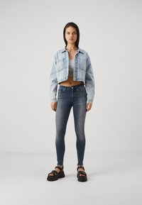 Svijetloplavi denim kratki kaput, sivi top bez rukava, tamnoplave uske traperice i crne platforme. Model stoji protiv neutralne pozadine.
