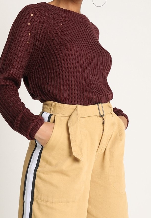 Femme portant un pull en maille bordeaux et un pantalon beige taille haute avec des bandes latérales blanches et noires, mains dans les poches.