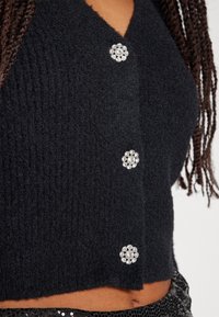 Černý pletený cardigan s výstřihem do V, ribaným textura a dekorativními kamínkovými knoflíky uspořádanými do květinového vzoru. Krátká délka.