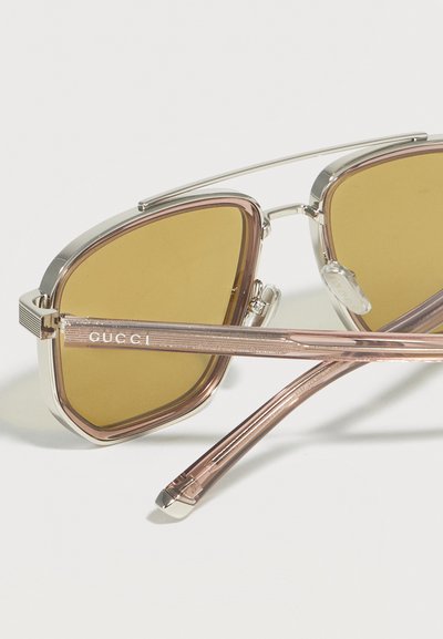 Γυαλιά ηλίου με μεταλλικό σκελετό, κίτρινους φακούς, ροζ βραχίονες και λογότυπο "GUCCI" στον βραχίονα, τοποθετημένα σε απλό λευκό φόντο.