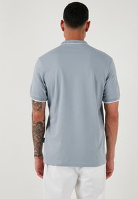 Světle modré polo tričko s uvolněným střihem, s klasickým límečkem a bílými akcenty na rukávech. Hladká textura, jednoduchý design.