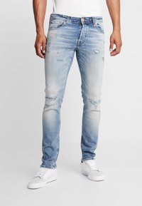 Jeans de mezclilla azul claro desgastados con un ajuste estrecho, que presentan áreas desteñidas y roturas visibles. Combinados con zapatillas blancas.