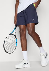 Ναυτικό μπλε τένις σορτς με αεραγωγούς, συνδυασμένα με λευκά αθλητικά παπούτσια και κρατώντας ένα μαύρο και μπλε ρακέτα τένις.