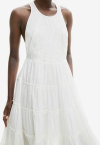 Vestido blanco sin mangas con un cuerpo ajustado y falda de volantes en capas. Presenta un sutil bordado en el cuerpo y una tela ligera.