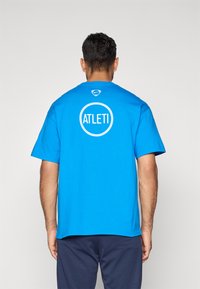 Mand med ryggen til iført en lysblå Nike T-shirt med "ATLETI" inde i en hvid cirkel på ryggen, kombineret med mørke bukser.