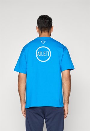 Man facing away wearing a bright blue Nike T-shirt with "ATLETI" inside a white circle on the back, paired with dark pants.