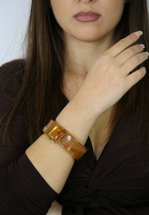 Bracelet en acrylique marron avec une fermeture rectangulaire ornée d'accent doré. Fini lisse, design large, porté sur un poignet nu.