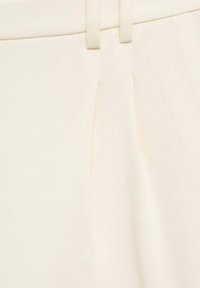 Gros plan d’un pantalon crème cintré avec une taille munie de passants de ceinture et un pli marqué à l’avant.