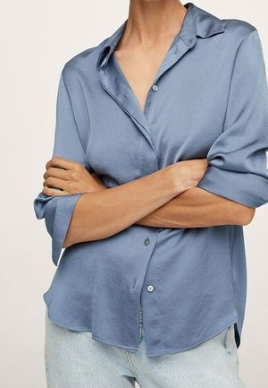 Kvinde iført en løs, blå skjorte med knapper og opkørte ærmer, armene krydsede, kombineret med lyseblå jeans.