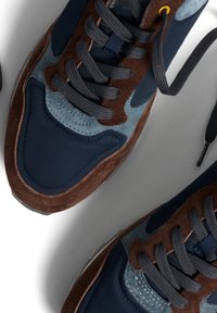 Zapatillas de color azul marino con acentos de ante marrón, que cuentan con un tejido texturizado, cordones gruesos y una suela acolchada. Se aprecian detalles de costura notables.