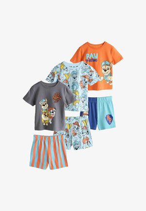 Bambino con una maglietta grigia con tre cuccioli animati con elmetti da cantiere e il testo "Tutti zampette dentro!" in piedi all'interno accanto a un cuscino a righe.