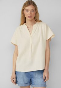 Hellbeige kurzärmlige Bluse aus strukturiertem Stoff mit mandarinfarbenem Kragen und Frontblende. Kombiniert mit Jeansshorts.