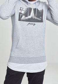 Felpa con cappuccio grigia con tasca a marsupio, con una grafica in bianco e nero di mani in preghiera e la parola "pray" in corsivo sotto.