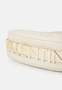 Kerek, beige bőr táska, amelyen a Valentino felirat található, és az elülső oldalon arany fémes betűkben olvasható a "VALENTINO" szó.