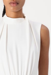 Robe blanche sans manches avec un col montant, tissu plissé et texture lisse, présentant des détails froncés à la taille.