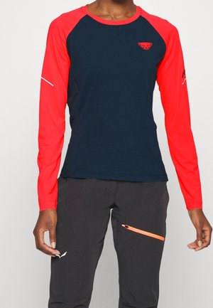 Person iført en marineblå og knaldrød sportsbluse med lange ærmer og sorte bukser med en orange lynlåslomme.