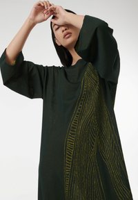 Groene oversized jurk van een gestructureerde stof, met een opvallend geel lijnpatroon aan één kant en brede mouwen.
