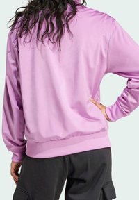 Person med långt hår som bär en glänsande ljuslila jacka och svarta byxor, stående med en hand på höften, vänd bort.