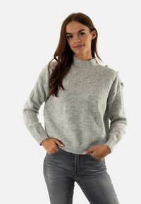 Maglione grigio chiaro a maglia con un motivo a diamante testurizzato, colletto alto e dettagli con bottoni sulla spalla, abbinato a jeans grigio scuro.