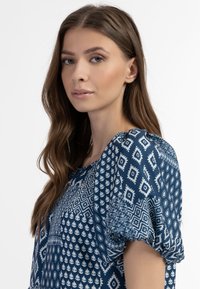 Blå mønstret bluse med korte puffærmer, geometriske og bladmotiver i hvid, lavet af et blødt stof med en glat tekstur.