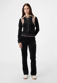 Sort sportstrøje med beige detaljer, lynlås foran og krave; kombineret med sorte bukser. Materialet virker blødt med en glat finish.