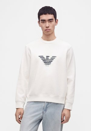 Jeune homme portant un sweat-shirt blanc avec un logo foncé et un jean bleu clair, debout devant un fond clair uni.