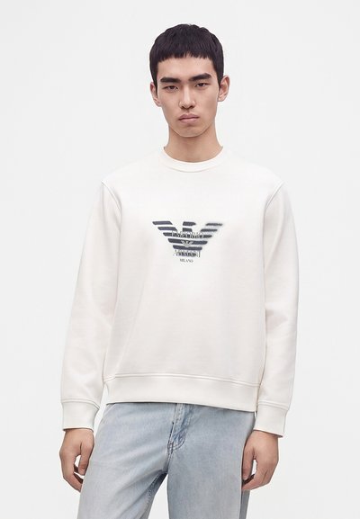 Jeune homme portant un sweat-shirt blanc avec un logo foncé et un jean bleu clair, debout devant un fond clair uni.