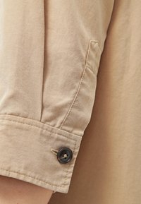 Poignet de chemise à manches longues beige avec un seul bouton foncé et des coutures visibles sur un tissu léger.
