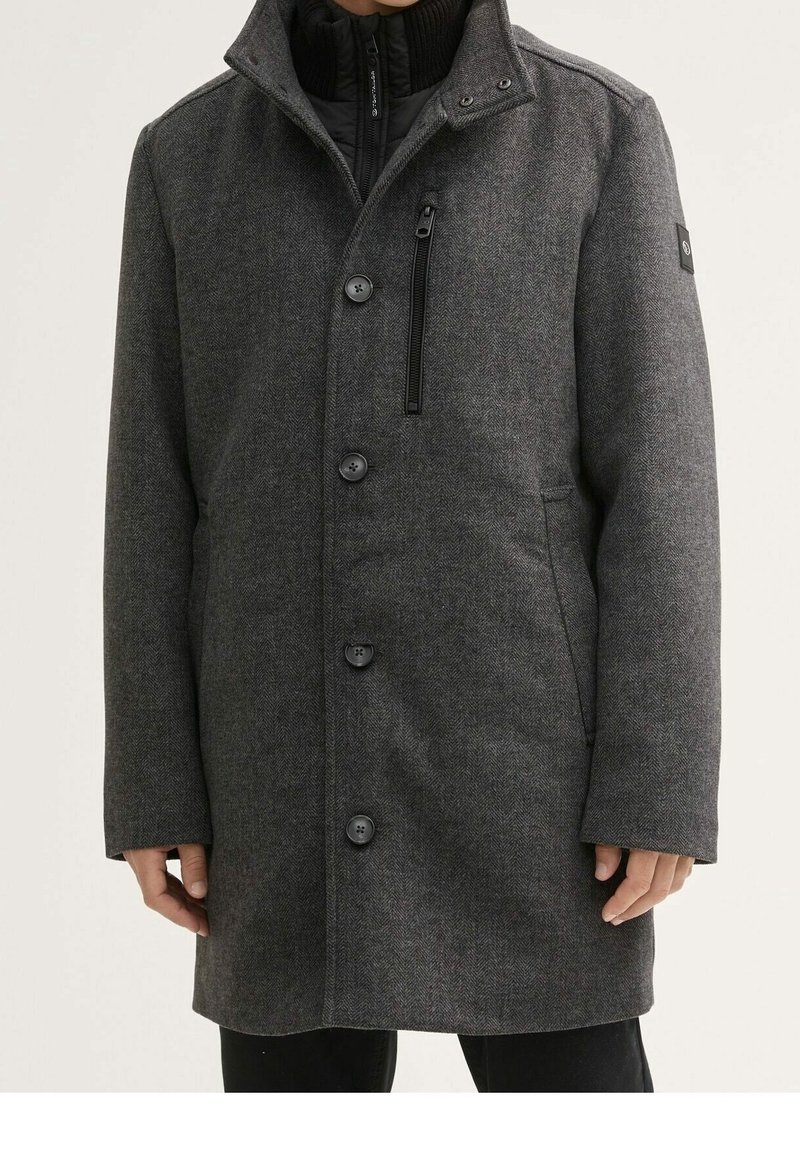 Manteau en laine gris foncé à chevrons avec fermeture à boutons, poche poitrine zippée, poches latérales et col montant, porté sur un pull noir zippé.