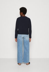 Maglietta a maniche lunghe blu navy abbinata a jeans a gamba larga di colore azzurro chiaro e scarpe nere slip-on, vista da dietro su uno sfondo semplice.