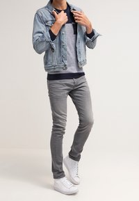 Jeansjacke mit hellem Waschton, grauer Rundhalspullover, graue Slim-Fit-Jeans und weiße Turnschuhe. Lässiges, geschichtetes Outfit ohne Muster oder Akzente.