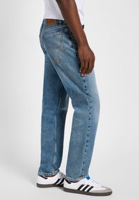 Jeans de mezclilla azul claro con un corte relajado, que cuentan con un parche de cuero en la cintura, diseño de cinco bolsillos y un desvanecimiento sutil. Llevados con zapatillas blancas.