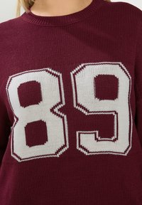 Suéter burdeos con una textura de punto grueso que presenta un gran número blanco "89" destacado en la parte frontal. Cuello redondo.