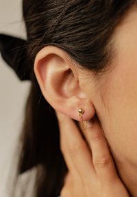 Goldene Steckerohrringe mit einer kleinen Kugel und einem zarten Kettendetail. Nahaufnahme des Ohrs, das glatte Haut und strukturiertes Haar zeigt.