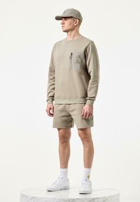 Beige sweatshirt med sidelomme, parret med beige shorts; hvide sneakers og sokker fuldender outfittet. Glat stof, afslappet design.