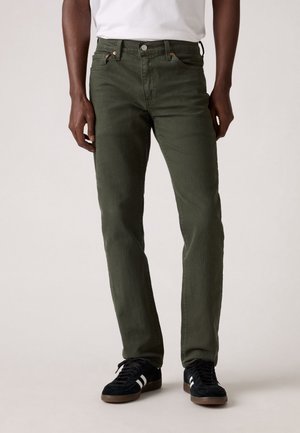 Mies, jolla on oliivinvihreät suoralahkeiset farkut, valkoinen t-paita ja mustat tennarit valkoisilla raidoilla, seisoo yksivärisen taustan edessä.