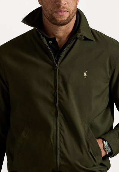 Veste zippée verte avec un col, présentant une finition texturée et un logo sur la poitrine gauche ; portée sur un polo noir.
