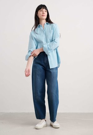 Chemise boutonnée rayée bleu clair avec poche poitrine et manches retroussées, associée à un jean large bleu foncé et des baskets blanches.