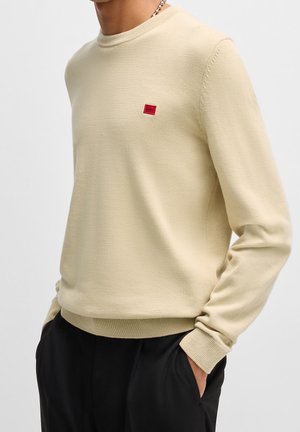 Mann trägt einen beigen Langarmpullover mit kleinem rotem Hugo-Logo auf der Brust und schwarze Hose, Hände in den Taschen, vor einfachem grauem Hintergrund.
