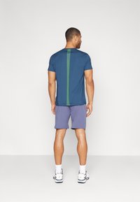 Tmavomodré športové tričko s vertikálnym zeleným akcentom, krátkymi rukávmi a voľným strihom, spárované so svetlofialovými šortkami a bielymi topánkami.