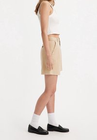 Pantalones cortos de algodón beige con un corte clásico, bolsillos frontales y una textura suave, combinados con mocasines negros y calcetines blancos.